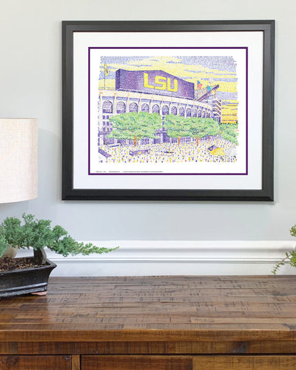  Framed LSU Tiger drawing of stadium in yellow and purple with green trees made of words on wall above wood desk. 