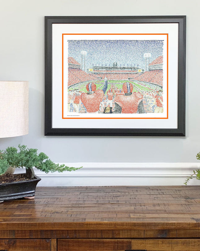 Matted and framed word art print of Clemson Memorial Stadium, one of the best Clemson gifts, hangs on wall over wood table.