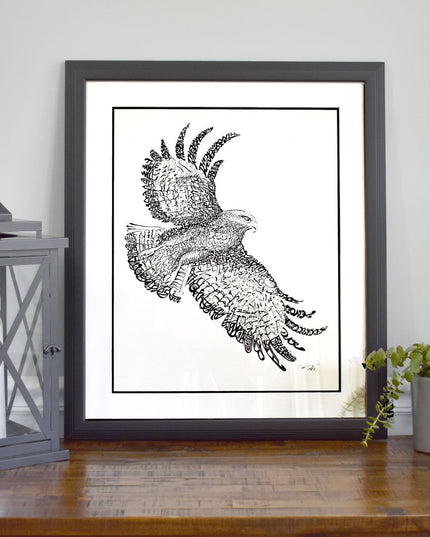 Framed art of St. Joseph Hawk with handwritten phrase "The Hawk Will Never Die" and white background on dresser by lantern.