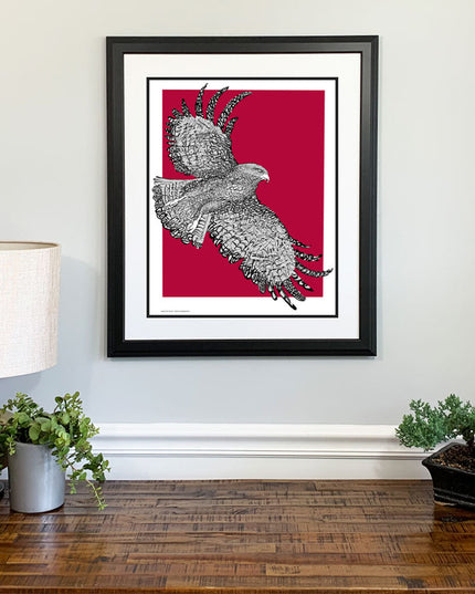 Framed art of St. Joseph Hawk with handwritten phrase "The Hawk Will Never Die" and red background on wall above dresser.